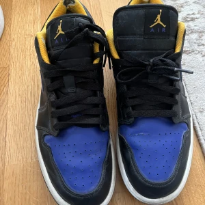 Nike Air Jordan 1 Low svart/blå/gul - Säljer ett par Nike Air Jordan 1 Low med svart läder, blå detaljer på tå och sidor samt gula insidor. Klassisk Jumpman-logga på plösen och AIR-tryck. Skorna har svart snörning och vit sula. Vid snabbt affär kan priset diskuteras.