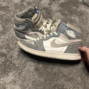Nike Air Jordan 1 High i blått och vitt - Säljer ett par Nike Air Jordan 1 High med vit bas och gråblå detaljer. Skorna har klassisk snörning, rund tå och hög siluett. Perfekta för dig som gillar streetwear och sneakers med ikonisk design.