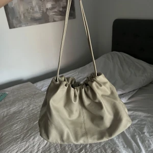 Beige handväska med dragsko - Säljer en beige handväska med dragskostängning och långa axelband. Väskan har en mjuk, slouchy form och är tillverkad i skinnliknande material. Perfekt för dig som gillar minimalistisk och stilren design.