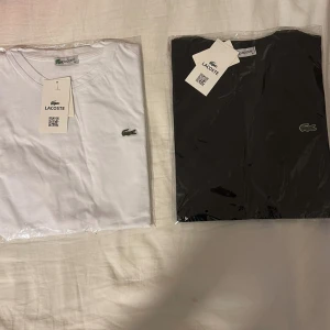 Lacoste t-shirt i vitt och svart - Två klassiska t-shirts från Lacoste, en vit och en svart, båda med den ikoniska krokodilloggan broderad på bröstet. T-shirtsen har rund hals och korta ärmar. Perfekt basplagg för garderoben.