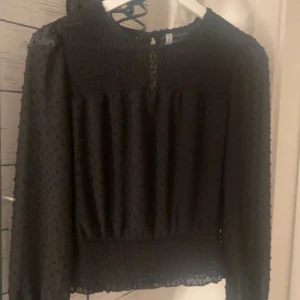 Svart blus med transparenta ärmar - Säljer en svart blus med långärmade, transparenta ärmar och diskret prickigt mönster. Blusen har smockdetaljer vid halsringning och midja samt knäppning bak. Perfekt för dig som gillar stilrena och trendiga plagg.