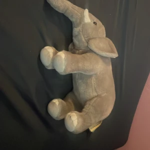 Mjukisdjur elefant - Säljer ett gulligt mjukisdjur i form av en elefant. Den är grå med vita detaljer på fötter och betar. Elefanten har stora öron, lång snabel och svarta ögon. Perfekt som gosedjur eller dekoration i rummet.