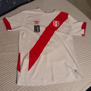 Peru landslagströja Umbro 17-18 säsongen  med nummer 10 Farfan - Vit Peru fotbollströja från Umbro med röd diagonal rand, nummer 10 och namnet Farfan på ryggen. Tröjan har korta ärmar och Perus landslagsemblem på bröstet. .