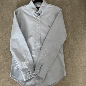 Ljusblå skjorta från Ralph Lauren - Klassisk ljusblå skjorta från Ralph Lauren i slim fit-modell. Skjortan har knappar framtill, lång ärm och den ikoniska broderade loggan på bröstet. Perfekt för en stilren och tidlös look.