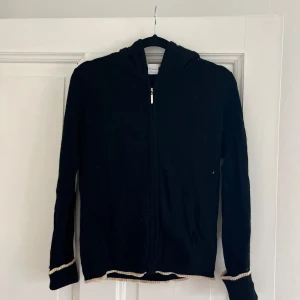 Svart cashmere hoodie med dragkedja  - Mysig svart hoodie i 100% cashmere kofta. Tröja med dragkedja framtill, huva och kontrastfärgade detaljer vid ärmslut och nederkant. Har ett litet hål på högra arm, något man kan fixa med lite tråd och en nål.