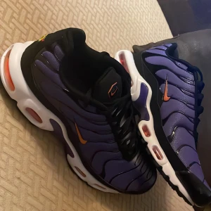 Nike Air Max Plus TN lila och svart - Nike Air Max Plus TN sneakers i lila och svart med orange swoosh och detaljer. Skorna har en vågig design på ovandelen, vit sula med synliga luftbubblor och svarta samt gula detaljer på undersidan. Perfekta för dig som vill sticka ut med färg och stil.