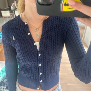 Mörkblå kabelstickad kofta från Polo Ralph Lauren - Säljer en klassisk mörkblå kabelstickad kofta från Polo Ralph Lauren med vita knappar och broderad logga på bröstet. Koftan har rund halsringning och långa ärmar. 