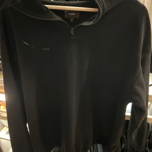 Svart fleece-tröja med dragkedja från LXA - Säljer en svart fleece-tröja från LXA i storlek L. Tröjan har halv dragkedja framtill och hög krage, med resår i midjan. Enkel och stilren design med liten LXA-logga på bröstet.