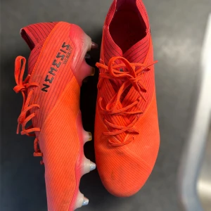 Röda Adidas Nemesis fotbollsskor - Säljer ett par röda Adidas Nemesis fotbollsskor med snörning och platt sula. Skorna har en modern design med tydliga linjer och är tillverkade i syntetmaterial. Perfekta för dig som vill sticka ut på planen!