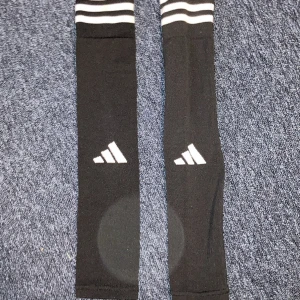 Svarta Slevves från Adidas - Säljer ett par svarta knästrumpor från Adidas med vita ränder upptill och vit Adidas-logga på framsidan. Perfekta för fotboll eller träning. Klassisk sportig look med bekväm passform.