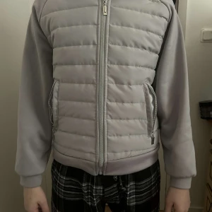 Ljusgrå pufferjacka med huva - Säljer en ljusgrå pufferjacka med huva och dragkedja framtill. Jackan har quiltad framsida och rygg, samt släta ärmar och två fickor med dragkedja. Perfekt för kyliga dagar och enkel att matcha med olika outfits.