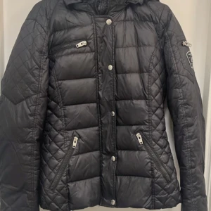 Svart quiltad jacka från Rockandblue - Säljer en svart quiltad jacka från Rockandblue med huva och dragkedjor på fickor och ärm. Jackan har snygga detaljer och en sportig look, perfekt för kyliga dagar. Märke på ärmen och tryckknappar framtill.