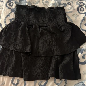 Svart volangkjol från Seaside - Säljer en svart kjol från Seaside med volangdetalj upptill och smockad midja. Kjolen är kort och har en luftig känsla, perfekt för varma dagar. Enkel att matcha med olika toppar för en trendig look.