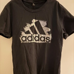 Svart t-shirt från Adidas med logga - Svart t-shirt från Adidas med stor vit logga framtill. Klassisk rund halsringning och korta ärmar. Tillverkad i mjuk bomull, perfekt för en sportig och avslappnad stil.