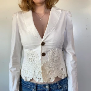 Vit kavaj med virkade detaljer från Moschino - Säljer en vit kavaj från Moschino med två knappar framtill och unika virkade spetsdetaljer längs nederkanten. Jackan har lång ärm och markerad krage, perfekt för dig som vill sticka ut med en cool och feminin stil.