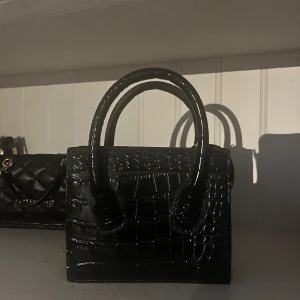 Svart handväska i krokopräglat mönster - Säljer en svart handväska med krokopräglat mönster och två stadiga handtag. Väskan har ett stilrent och elegant utseende med ett rymligt huvudfack och innerficka med dragkedja. Perfekt för dig som gillar trendiga accessoarer.