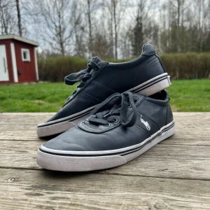 Mörkblå sneakers från Polo Ralph Lauren - Säljer ett par svarta sneakers från Polo Ralph Lauren med vit logga på sidan och vita sulor. Skorna har snörning och är tillverkade i skinn med klassisk låg profil. Perfekta för en stilren och avslappnad look. Högerskon har en fläck på fronten men annars är dom i bra skick.