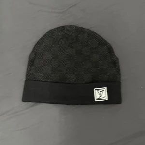 Svart mössa med logotyp L*V - Snygg svart mössa med diskret rutigt mönster och en broderad logotyp framtill. Perfekt för kyliga dagar.