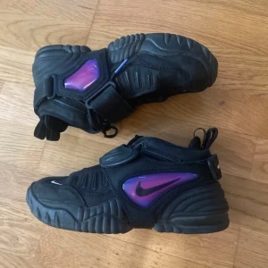 Nike x Ambush sneakers med lila detaljer - Säljer ett par svarta Nike x Ambush sneakers med lila och vita detaljer. Skorna har en chunky sula, snörning och tydliga Nike-loggor på sidorna samt Ambush-tryck på hälen. Perfekta för dig som gillar unika och sportiga sneakers. Väldigt gott skick använda ett fåtal gånger 💜