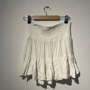 Vit volangkjol med smock - Söt vit kjol med smock upptill och volang nertill. Kjolen är kort och har en luftig känsla, perfekt för varma dagar. Passar till många olika stilar och är lätt att matcha med olika toppar.