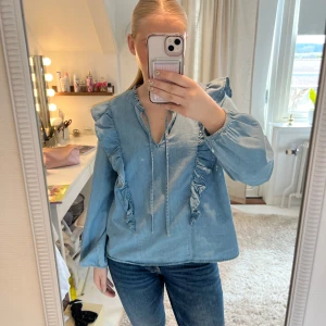 Blå jeansblus med volang från Kappahl  - Säljer en ljusblå ”jeansblus” (100% bomull) från kappahl med volangdetaljer längs axlar och bröst. Blusen har v-ringning med knytband och långa ärmar. Köpt för 449 kr! Använd 1 gång. Som ny!❤️