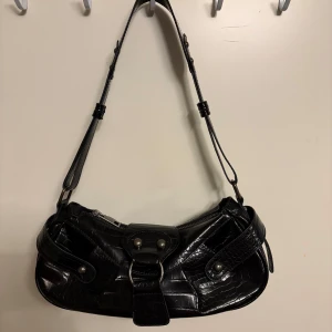 Svart axelväska i croco-mönstrat skinn från Urban Outfitters - Snygg svart axelväska från Urban Outfitters i croco-mönstrat skinnimitation. Väskan har justerbart axelband, dragkedja och spänndetaljer framtill. Perfekt för att lyfta din outfit med en trendig accessoar.