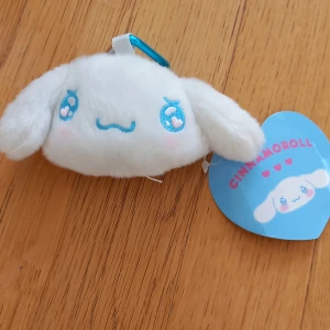Vit Cinnamoroll plånbok med hjärtformat spänne - Supersöt liten plånbok i form av Cinnamoroll från Sanrio. Den är vit, mjuk och fluffig med broderade blå detaljer och rosa hjärtan på kinderna. Plånboken har ett hjärtformat blått spänne och ett fack med ljusblått foder.