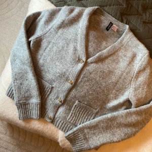 Ljusgrå kofta - Mysig ljusgrå kofta från H&M med snygga guldiga knappar och ribbade detaljer på fickorna och nederkanten. Koftan har långa ärmar och är stickad i ett mjukt material. Inga defekter och näst intill helt oanvänd. Skriv för fler bilder och vid frågor☺️