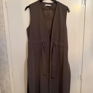 Khaki lång väst med knytband från Mango - Säljer en stilren brun väst från Mango med långa knytband i midjan. Västen är ärmlös, har en rak passform och är perfekt för lager-på-lager-stil. Knappt använd, passar även storlek L 