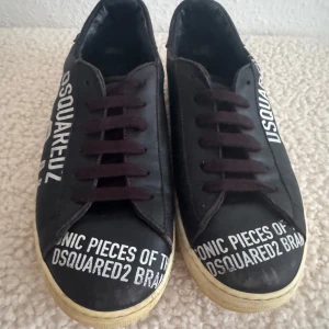 Svarta sneakers från Dsquared2 strl 37 - Svarta sneakers från Dsquared2 med vit text på sidan och framtill. Skorna har svarta snören, vit sula och är tillverkade i skinn. Snygg streetstil med tydlig branding och klassisk rund tå. Skavanker framme på skorna och sulan är gulaktig.