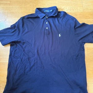 Marinblå pikétröja från Ralph Lauren - Klassisk marinblå pikétröja från Polo Ralph Lauren i bomull. Tröjan har korta ärmar, krage och tre knappar framtill. Ikoniska broderade loggan i vitt på bröstet. Perfekt för en clean och stilren look.