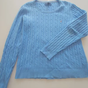 Ljusblå kabelstickad tröja Gant  - Kabelstickad ljusblå tröja från Gant i bomull med rund halsringning och broderad logga på bröstet. Tröjan har ribbade muddar och är långärmad, perfekt för en chill och stilren look.