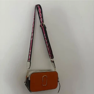 Marc Jacobs axelväska med logoband - Snygg axelväska från Marc Jacobs i brunt, vitt och svart skinn. Väskan har två dragkedjor upptill och en bred axelrem med rosa och svart logotyp. Perfekt storlek för det viktigaste och en stilren metallogo framtill.