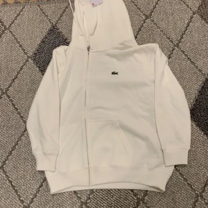 Vit hoodie från Lacoste med dragkedja - Snygg vit hoodie från Lacoste med klassisk krokodil-logga på bröstet. Tröjan har huva, dragkedja framtill och två fickor. Perfekt för en clean och sportig look. Tillverkad i mjuk bomull för extra komfort.