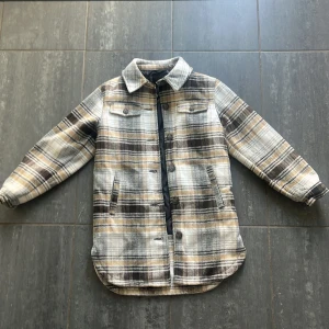 Rutig overshirt från Name It - Riktigt snygg rutig overshirt från Name It i beige, brunt och grått. Jackan har klassisk krage, knappar framtill och två bröstfickor med lock. Fodrad insida för extra värme och långa ärmar med knapp vid ärmslut. 