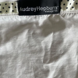 Vit skjorta Audrey Hepburn Vintage 55 - Vit skjorta med stora fickor framtill och snygga beige knappar. Insidan av knappslån har ett band i satin med svarta prickar som ger en cool retrovibe. Skjortan är i bomull och har en klassisk, rak passform. Perfekt för dig som gillar detaljer och vintagekänsla.