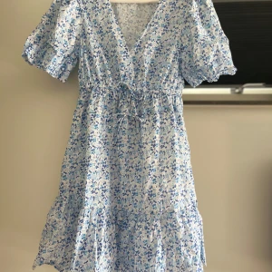Blommig kortklänning från C XS - Supersöt kortklänning med blått blommönster  från C. Klänningen har v-ringning, puffiga kortärmar och knyt i midjan. Perfekt för sommarens alla tillfällen och midsommar och har en luftig, flowy känsla. Säljer den på grund av att den är för liten för mig. Klänningen är i perfekt skick!