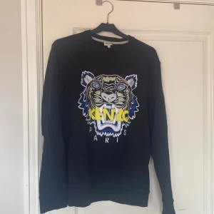 Svart Kenzo tröja  - Svart tröja från Kenzo med ikoniskt broderat tigerhuvud i blått, gult och vitt framtill. Rund halsringning och långa ärmar. Perfekt statement-piece för dig som vill sticka ut med en cool design och snygga färgdetaljer.