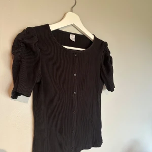 Svart ribbad topp med puffärm  - Svart ribbad topp från Lindex med korta puffärmar. Toppen har knappar framtill och är i mjukt och jätte skönt material. Perfekt för dig som gillar en enkel men ändå trendig look. Storlek 146/152 men ganska stor i storlek då den inte är tvättad eller använd så mycket. Den passar fortfarande mig som har xs/s. 💖💖