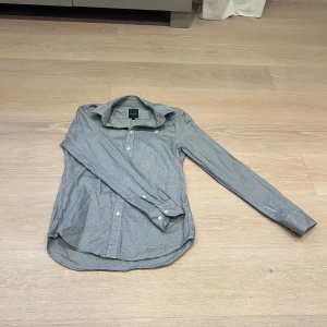 Grå skjorta från G-Star Raw, M - Snygg grå skjorta från G-Star Raw i storlek M. Skjortan har ett diskret rutigt mönster, klassisk krage och vita knappar framtill. Långärmad modell, perfekt för en clean och stilren look.