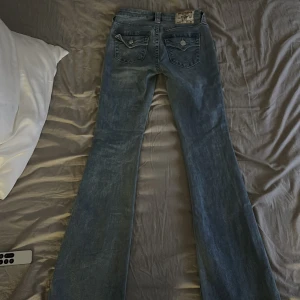 True Religion Becca Bootcut Jeans ljusblå - Säljer ett par True Religion Becca Mid Rise Bootcut jeans i ljusblå tvätt. Snygg passform med klassiska bakfickor och silverfärgade knappar. Jeansen har bootcut-ben och är tillverkade i mjukt denim. Perfekta för dig som gillar en tidlös och trendig look. Midje mått 81, innrebenslängd 83. Frakt betalas av köpare 