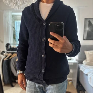 Mörkblå kofta från Hampton Republic - Säljer en mörkblå kofta från Hampton Republic med grovstickad struktur, sjalkrage och knappar framtill. Snygga mockadetaljer på armbågarna och ribbade muddar. Perfekt för lager-på-lager och chillare dagar.