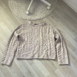 Beige stickad tröja från 157 - Mysig beige stickad tröja från 157 med grovt flätmönster och rund hals. Tröjan har lång ärm och en lite boxig, avslappnad passform. Perfekt för lager-på-lager och chill dagar. Passar dig som gillar en enkel men ändå snygg look.