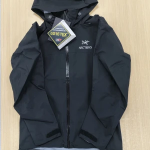 Svart Arc'teryx vindjacka Gore-Tex S - Svart vindjacka från Arc'teryx i storlek S, tillverkad i slitstark Gore-Tex med minimalistisk design. Jackan har huva, hel dragkedja framtill och justerbara ärmslut med kardborre. Perfekt för dig som vill ha en lätt och smidig jacka till vardagsäventyr.