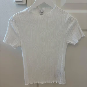 Vit topp ribbad - Säljer en vit ribbad topp från New Yorker med korta ärmar och vågiga kanter. Toppen har en lätt stretchig känsla och ett snyggt plisserat mönster som ger en cool struktur. Aldrig använt.