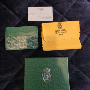 Grön Goyaard plånbok med mönster - Säljer en grön plånbok från Goyaard med deras ikoniska mönster i vitt och beige. Plånboken är i skinn och har flera kortfack. Kommer med original ask, tygpåse och äkthetskort. Perfekt för dig som vill ha något exklusivt och stilrent i fickan.