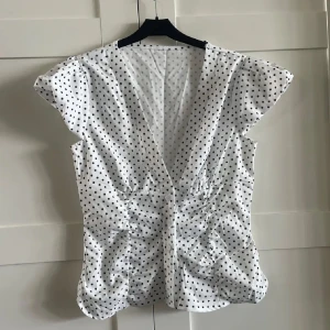 Vit prickig blus med v-ringning - Supersöt vit blus med svarta prickar och djup v-ringning. Korta ärmar och smickrande rynkade sidor ger en snygg siluett. Knäppning framtill och lätt figurnära passform. 