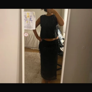 Svart långklänning med paljetter - Säljer en svart tvådelad långklänning med glittriga paljetter och fransdetaljer. Toppen är ärmlös och har rund halsringning, kjolen är rak och hellång. Perfekt för dig som vill sticka ut med en glamorös look. Med prislapp 