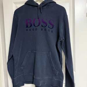 Mörkblå hoodie från Hugo Boss - Snygg mörkblå hoodie från Hugo Boss med stor lila BOSS-logga på bröstet. Klassisk känguruficka framtill, huva som är vattentät  med snörning och långa ärmar. Perfekt för dig som gillar stilrena plagg.