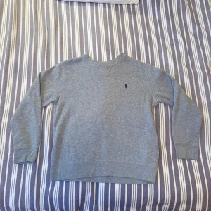 Grå sweatshirt från Polo Ralph Lauren - Klassisk grå sweatshirt från Polo Ralph Lauren med broderad marinblå logga på bröstet. Tröjan har rund halsringning, långa ärmar och är tillverkad i mjuk bomullsmix. Passar riktigt bra till de flesta klädesplagg och året runt, perfekt för en chill och stilren look. Skick: 9/10. Hör gärna av dig vid intresse om fler bilder eller frågor😊